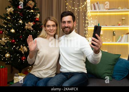 Porträt eines glücklichen weihnachtspaares mit Smartphone zur Aufnahme von Online-Grußvideos, Ehemann und Ehefrau blicken auf die Kamera, um das neue Jahr zu feiern und sitzen auf dem Sofa im Wohnzimmer. Stockfoto