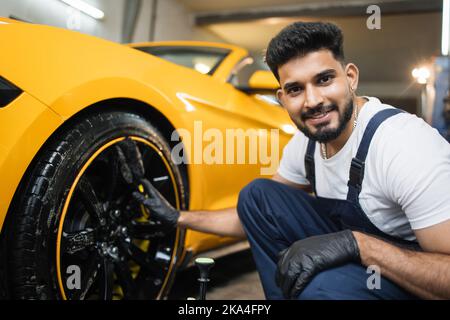 Professionelle männliche Arbeiter in Overalls und schwarzen Schutzhandschuhen, Reinigung von Leichtmetallfelgen Felgen von Luxuswagen mit einer speziellen Bürste für Gussräder in einer Detaillierungswerkstatt. Stockfoto