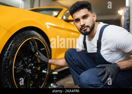 Professionelle männliche Arbeiter in Overalls und schwarzen Schutzhandschuhen, Reinigung von Leichtmetallfelgen Felgen von Luxuswagen mit einer speziellen Bürste für Gussräder in einer Detaillierungswerkstatt. Stockfoto