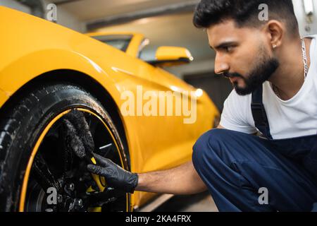 Professionelle männliche Arbeiter in Overalls und schwarzen Schutzhandschuhen, Reinigung von Leichtmetallfelgen Felgen von Luxuswagen mit einer speziellen Bürste für Gussräder in einer Detaillierungswerkstatt. Stockfoto