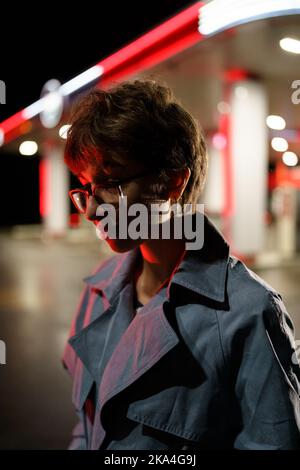 Ruhige, traurige junge Frau, die abends vor dem Hintergrund von farbigen Neonlichtern auf der Stadtstraße steht. Stockfoto