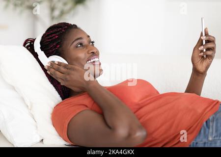 Portrait Von Happy Black Female Trägt Kopfhörer Entspannung Mit Smartphone Zu Hause Stockfoto