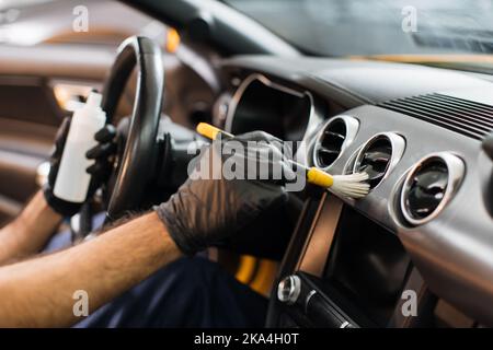 Beschnittenes Bild von Händen eines jungen männlichen Arbeiters in schwarzen Schutzhandschuhen, Reinigung des Armaturenbretts mit weicher Bürste und spezieller antistatischer Kunststoff-Ziermilch. Stockfoto