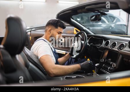 Attraktiver bärtiger Autodienstmitarbeiter in Handschuhen, weißem T-Shirt und Overalls, der mit einer speziellen weichen Bürste Staub von der Kunststoffplatte des Luxuswagens reinigt. Auto Reinigung und Detaillierung Konzept, Trockenwäsche. Stockfoto