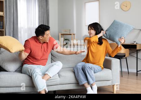 Asiatische Ehegatten im Homewear sitzen auf der Couch und haben Kissenschlacht Stockfoto