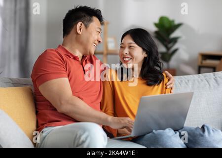 Glückliche chinesische Ehepartner, die sich zu Hause mit einem Laptop verbinden Stockfoto