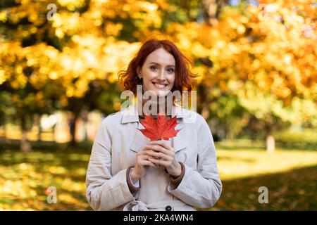 Porträt einer lächelnden, hübschen jungen europäischen rothaarigen Dame im Regenmantel halten im Herbst im Stadtpark ein rotes Blatt Stockfoto