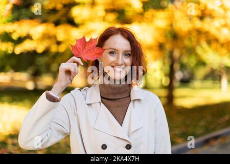 Fröhliche, hübsche junge, rothaarige Frau aus europa im Regenmantel hält ein rotes Blatt und schaut in den Park in die Kamera Stockfoto