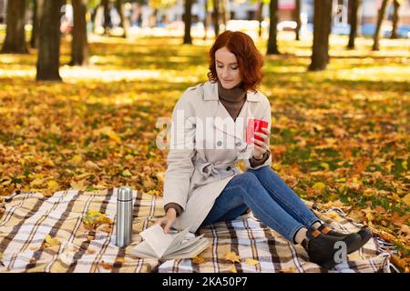 Serious smart junge europäische rothaarige Dame im Regenmantel genießen Tasse heißen Getränk aus Thermoskannen, Buch lesen Stockfoto