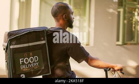 Restaurantlieferant geht zum Eingang des Bürogebäudes, junger Mann arbeitet als Lebensmittelkurier auf dem Fahrrad. afroamerikanische Lieferperson mit Thermosack, die Fahrrad auf der Straße reitet. Stockfoto