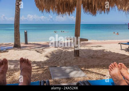Blick auf die Füße von Mann und Frau auf den Sonnenliegen am Sandstrand. Wunderschöne türkisfarbene Wasseroberfläche des Atlantischen Ozeans im Hintergrund. Aruba. Stockfoto