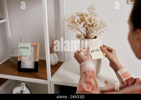 Selektiver Fokus auf Hände von jungen Frauen, die Notizpapier mit neuem spanischen Wort auf weiße Vase mit getrockneten Blumen auf dem Regal kleben Stockfoto