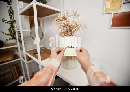 Hände einer jungen Frau kleben Notizpapier mit neuen spanischen Wort auf weiße Porzellanvase stehen auf dem Regal an der Wand des Schlafzimmers Stockfoto