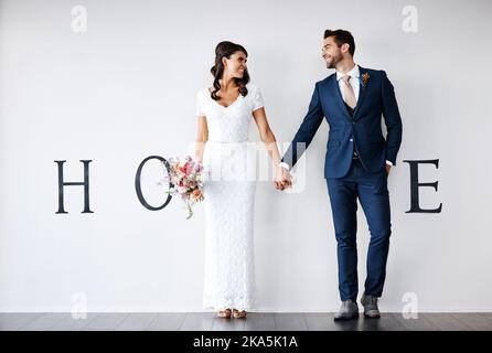 Lasst uns nach Hause gehen, Honig. Konzept-Studio-Aufnahme einer Braut und eines Bräutigams, die ein M im Wort Haus gegen eine Wand machen. Stockfoto