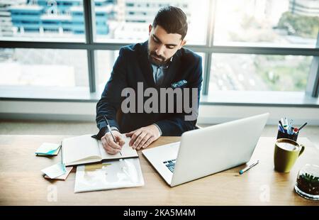 Ein junger Geschäftsmann, der in einem modernen Büro an seinem Schreibtisch in einem Notizbuch schreibt, klopft diese Arbeitsaufgaben aus dem Park. Stockfoto