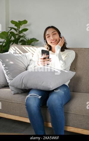 Ruhige und entspannte junge asiatische Frau, die lächelt, die Augen geschlossen, genießt es, Musik über ihre Kopfhörer zu hören, während sie auf der bequemen Couch in ihrem Leben chillt Stockfoto