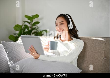 Attraktive junge asiatische Frau, die Kopfhörer trägt, genießt es, Filme oder Videos auf ihrem Tablet zu sehen, während sie morgens auf ihrer Couch im Wohnzimmer Kaffee schlürft Stockfoto