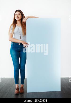 Es ist etwas, an dem sie steht. Studioaufnahme einer attraktiven jungen Frau, die sich vor weißem Hintergrund auf einem leeren Plakat lehnt. Stockfoto