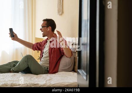 Ermutigt kaukasischen Mann mit Smartphone sitzt auf dem Bett im Schlafzimmer und emotional winkt Hand Stockfoto