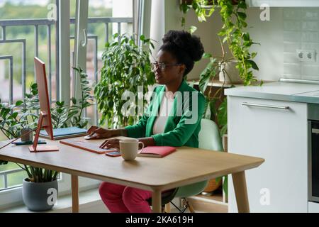 Junge afrikanische Geschäftsfrau in Brillen und formeller Kleidung, die aus der Ferne in einer gemütlichen Wohnküche arbeitet Stockfoto