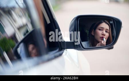 Junge Frau, die Lippenstift anwendet, und die Spiegelung im Autospiegel betrachtet. Stockfoto