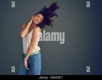 Lassen Sie Ihre Haare locker. Studioaufnahme einer unbeschwerten jungen Frau, die ihr Gesicht hält, während ihr Haar vor dunklem Hintergrund vom Wind geblasen wird. Stockfoto