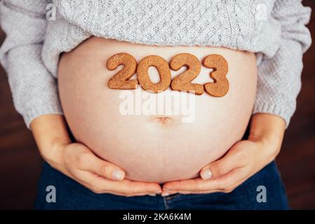 Zugeschnittenes Foto von einer Schwangeren, die Nummer 2023 von Ingwer-Cookies auf dem Bauch hält. Menschen und Erwartung Konzept. Glückliche Mutterschaft, gesundes Leben. Erwarten Stockfoto