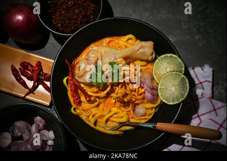 Blick von oben, Khao Soi Kai oder einzigartige nordthailändische Kursuppe mit Eiernudeln und Huhn auf einem schwarzen Esstisch. Thailändisches Essenskonzept Stockfoto