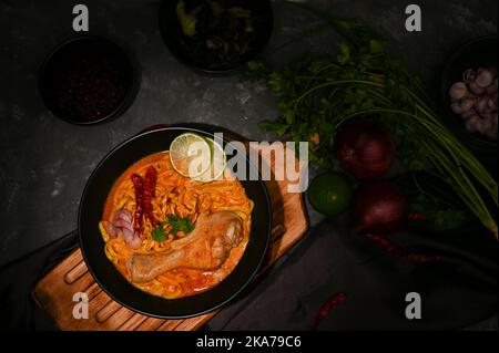 Overhead Shot, Khao Soi Kai oder cremiges und leicht würziges gelbes Kerri mit Hähnchendrumstick und Gemüse auf schwarzem Esstisch. Thailändisches Restaurant cui Stockfoto