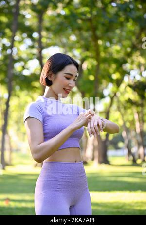 Portrait, schön fit junge asiatische Frau in Sportkleidung, die ihre Herzfrequenz auf ihrer smarten Uhr nach dem Joggen im Park überprüft. Gesundheit und Technologie c Stockfoto