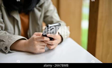Junge asiatische Frau in legerer Kleidung tippen auf ihr Telefon, chatten oder senden Nachrichten an ihre Freunde, während sie sich im Café. Zugeschnittenes Bild Stockfoto