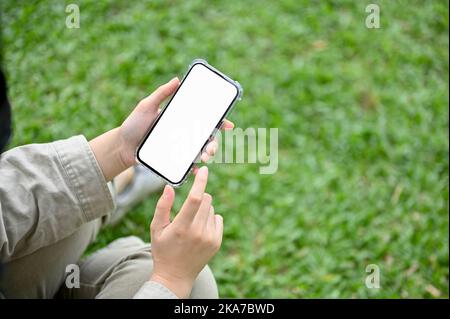 Draufsicht, Ein modernes Smartphone weißen Bildschirm Mockup ist in der Hand einer Frau über verschwommenem Hintergrund von grünem Gras. Junge asiatische weibliche sitzen auf Gras und Stockfoto