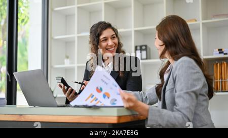 Eine glückliche und schöne kaukasische Geschäftsfrau aus der tausendjährigen Generation unterhält sich gerne mit ihrem Kollegen, während sie im Büro zusammenarbeitet. Stockfoto