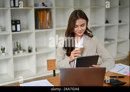 Wunderschöne und erfolgreiche junge kaukasische Geschäftsfrau oder weibliche Geschäftsführerin, die ihren Morgenkaffee schlürft und ihren Terminplan auf ihrem digitalen Tisch überprüft Stockfoto