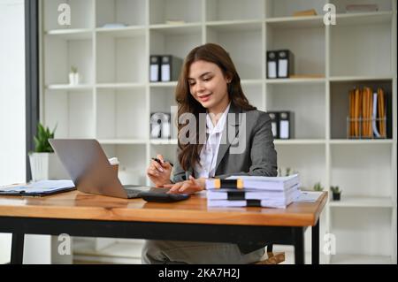 Professionelle und attraktive junge kaukasische Geschäftsfrau oder Buchhalterin mit Taschenrechner, Überprüfung ihrer finanziellen Verkaufsdaten, arbeiten bei ihrem d Stockfoto
