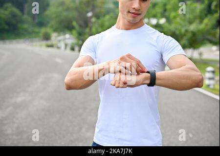 Hübscher und athletischer junger asiatischer Mann in Sportkleidung, der seine Smartwatch anschaut und auf seiner Sportuhr das Kalorienzählprogramm einstellt. Zugeschnittenes Bild Stockfoto