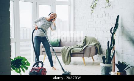 Die schöne junge Frau saugt zu Hause mit einem modernen Staubsauger auf den Boden und hört mit Kopfhörern Musik, tanzt und singt. Hausarbeit und Technikkonzept. Stockfoto