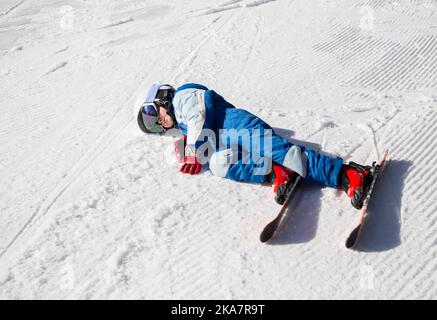 Müdes Kind in Helm, Brille, Ski und Winteroverall liegt auf dem Schnee. Fiel in den Schnee, hielt inne, um sich auszuruhen. Winterspaß. Kleiner Junge lernt Skifahren Stockfoto