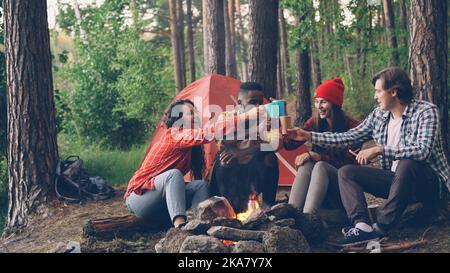 Sorglose Touristen toasten, klinken Tassen und Gläser mit heißen Getränken und trinken dann am Feuer im Wald in der Nähe des Zeltes. Freundschaft, Erholung und Jugendkonzept. Stockfoto