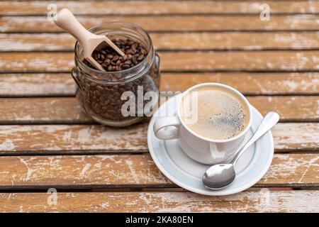 Kaffeetasse mit Glasgefäß, gefüllt mit Kaffeebohnen auf einem hölzernen Gartentisch. Stockfoto