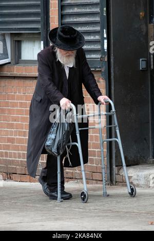 Ein älterer chassidischer Jude kehrt mit Hilfe eines Wanderers vom Einkaufen zurück. In Williamsburg, Brooklyn, New York City. Stockfoto