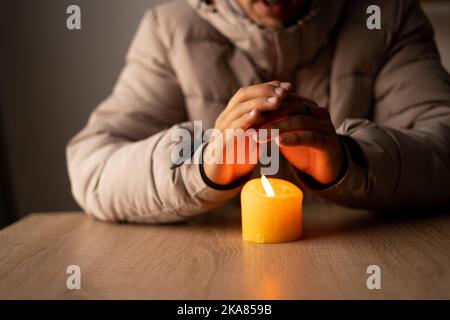 Ein Mann wärmt seine Hände aus der Nähe von einer brennenden Kerze im kalten Haus. Abschaltung von Heizung und Strom, Stromausfall, Stromausfall, Energiekrise. Stockfoto