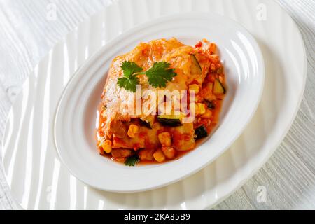 Nahaufnahme eines vegetarischen Enchilada-Auflaufes mit Tortillas, Sauce, Pinto-Bohnen, Mais, Zucchini und mexikanischem Käse auf weißem Teller Stockfoto