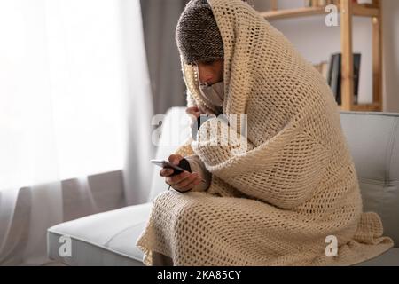 Unwell Mann Mieter in Decke sitzen in kalten Wohnzimmer Handy verwenden.leiden unter Mangel an Wärme und Strom. Ungesunder junger Mann kämpft gegen Kälte Stockfoto