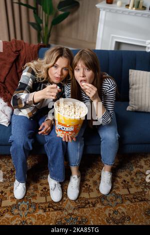 Zwei niedliche Frauen unterschiedlichen Alters konzentrieren sich darauf, Popcorn aus einem großen Eimer fernzusehen Stockfoto