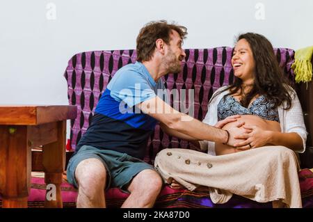 Ein Paar aus lateinamerikanischen Schwangeren eines kaukasischen argentinischen Mannes mit seiner brasilianischen Frau sitzt zu Hause auf dem Sofa und lächelt aufgeregt und fühlt den m Stockfoto