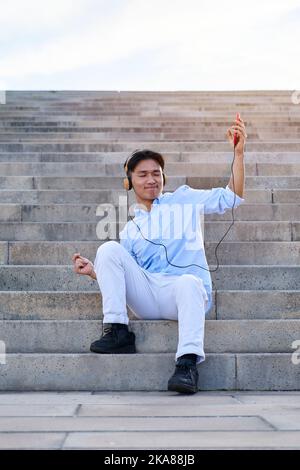 Asiatischer junger Mann, inspiriert vom Gefühl der Musik im Freien, während er auf der Treppe sitzt. Stockfoto