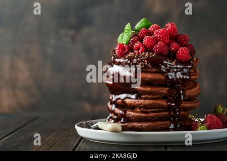 Schokoladenpfannkuchen. Pfannkuchen mit frischer Himbeere mit Schokoladenglasur oder Toppings in grauer Schale auf altem Holztischhintergrund. Hausgemachter Klassiker ameri Stockfoto