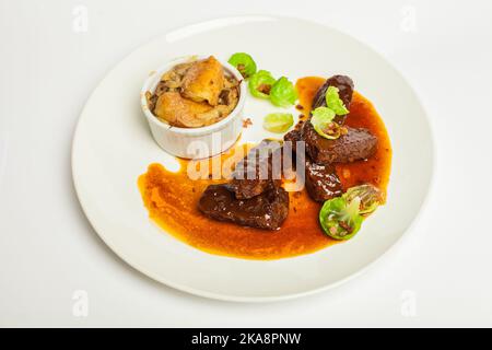 Leckere Kalbsbacken in roter Sauce mit grünem. Nahaufnahme auf einer weißen Platte auf weißem Hintergrund Stockfoto