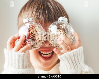 Kind mit dekorativen Kugeln für Weihnachtsbaum.Junge in Zopf-Strick übergroßen Pullover.gemütliches Outfit für kuscheln Wetter.Transparente Kugeln mit Spangeln. Stockfoto